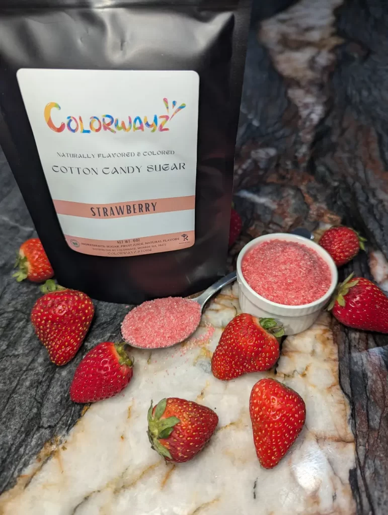 A bag of Colorwayz Strawberry Cotton Candy Sugar surrounded by fresh strawberries and a spoonful of pink sugar, showcasing natural ingredients and vibrant color.