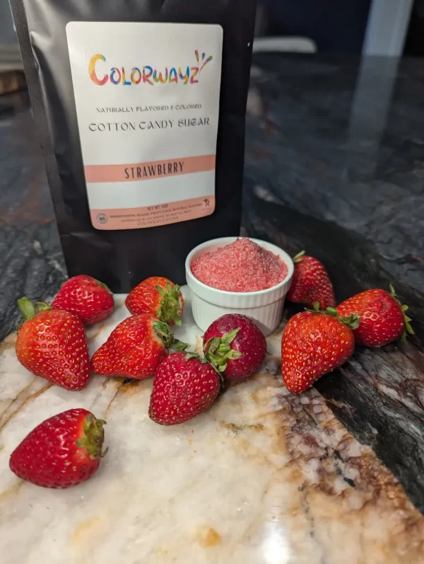 A bag of Colorwayz Strawberry Cotton Candy Sugar alongside a bowl of pink sugar and fresh strawberries, showing the natural, colorful ingredients.